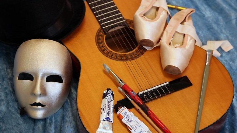 Teatermask, gitarr, målarpenslar och dansskor placerade på ett blått tyg